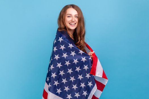 Portrait of satisfied woman wearing striped casual style sweater, standing wrapped in american flag, celebrating National independence day - 4th july. Indoor studio shot isolated on blue background.
