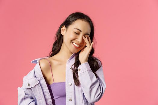 Lifestyle, people and emotions concept. Close-up portrait of cheerful attractive, stylish korean girl laughing, touching eye and smiling with closed eyes, having funny conversation, pink background.