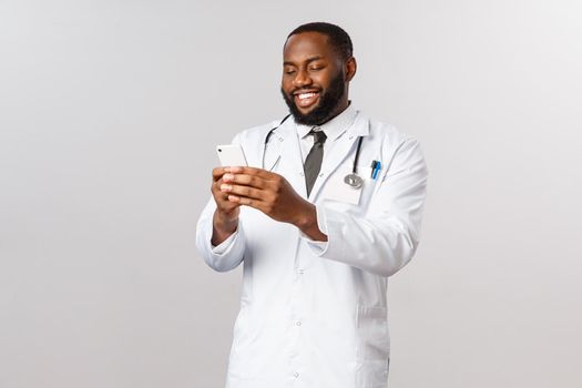 Covid19, pandemic and online medicine concept. Happy satisfied african-american doctor working hard in hospital during outbreak coronavirus, look at mobile phone, videcalling family with smartphone.