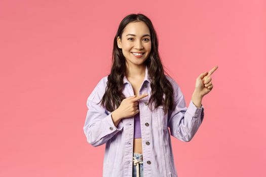 Portrait of cheerful asian girl, female student inviting people follow page, subscribe to page or visit link, pointing fingers right and smiling camera, standing happy over pink background.