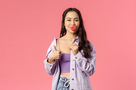 Holidays, lifestyle and people concept. Portrait of enthusiastic good-looking young woman in stylish outfit, asian girl show thumbs-up squinting sassy and holding big lips on stick.