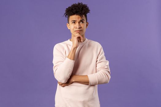 Portrait of complicated, young thoughtful man with dreads, look troubled up, thinking what to do, standing indecisive, grimacing not knowing answer, facing hard choices, purple background.