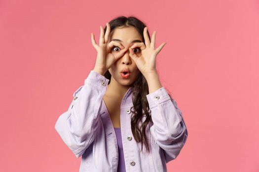 Close-up portrait of amused, speechless and impressed asian young girl seeing something curious and amazing, make glasses with fingers over eyes, fold lips say wow astonished, pink background.