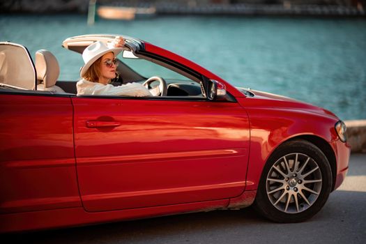 Outdoor summer portrait of stylish blonde woman driving red car convertible. Fashionable attractive woman with blond hair in a white hat in a red car. Sunny bright colors taken outdoors against the sea