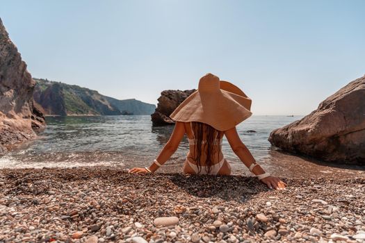 Tanned middle-aged woman with long hair and a white bathing suit. He sits on the seashore in a large sun hat with his back and looks at the sea