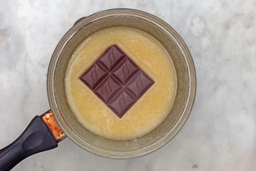 Top view of bain-marie with honey batter and chocolate in glass bowl on saucepan as ingredients of chocolate cake medovik on marble background