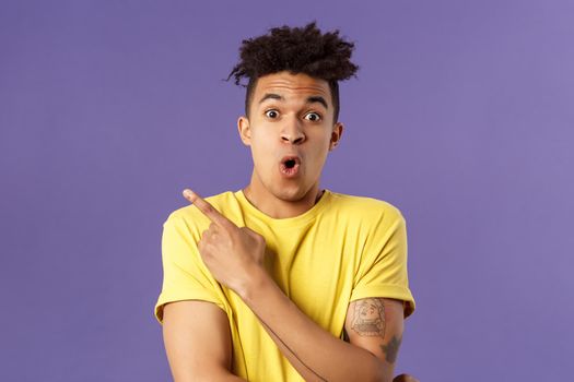 Portrait of surprised, excited young man showing cool new thing, pointing upper left corner, gasping, folding lips, stare camera impressed say wow, standing purple background.