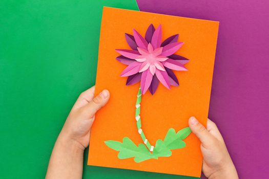 Top view of child hands holding papercraft violet crysanthemum flower made by child on orange worksheet on violet and green background. Present for mother s day