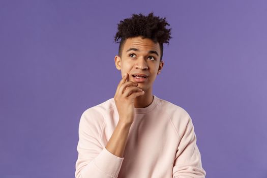 Waist-up portrait of thoughtful unsure young hispanic male student trying solve exercise, looking up thinking, pondering and calculating in mind, standing focused and indecisive purple background.