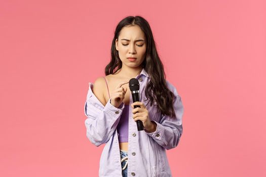 Portrait of awkward silly asian girl tap microphone as checking is it working, her turn to sing song on karaoke party, frowning complicated, standing pink background unsure how this thing works.