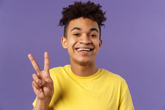 Close-up portrait of handsome upbeat young teenage guy with afro hairstyle, show peace sign and smiling, wear yellow t-shirt, staying optimistic and positive, purple background.