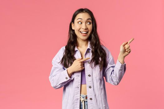 Portrait of curious, interested pretty asian woman in stylish outfit, pointing looking upper right corner with amused smile, see cool product, online courses or internet shopping advertisement.