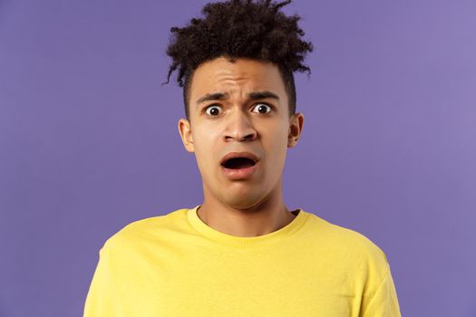 Close-up portrait of shocked, alarmed young man gasping, open mouth scared and frightened, staring camera concerned, facing troublesome shocking news, purple background. Copy space