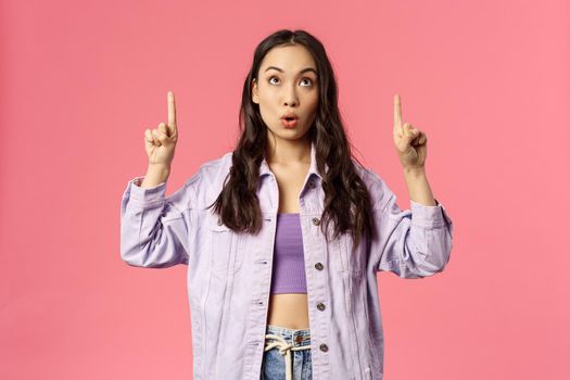 Portrait of curious and surprised young interested woman, mixed-race girl peeking and pointing fingers up, say wow, reading sign, visit interesting lecture or event, standing pink background.