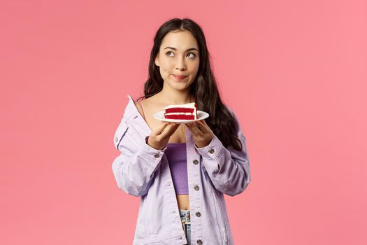 Troubled young asian girl wants to eat delicious piece cake but having doubts, hesitating of lots calories, smirk concerned look upper left corner thinking, making decision, pink background.