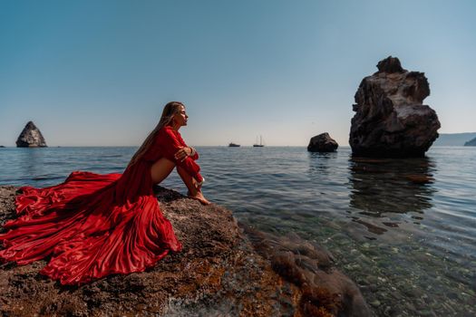 Beautiful sensual woman in a flying red dress and long hair, sitting on a rock above the beautiful sea in a large bay