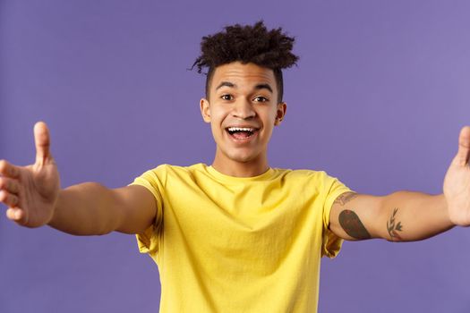 Close-up portrait of friendly cheerful young hispanic hipster guy giving warm welcome to dearest guests, spread hands sideways for greeting, cuddling or embracing person, smiling pleased.