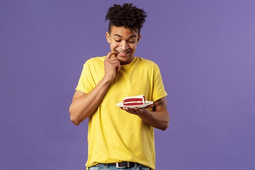 Celebration, party and holidays concept. Portrait of boyfriend cant resist temptation to eat last piece of cake, biting lips and smiling eager to have bite of dessert, hesitating, purple background.