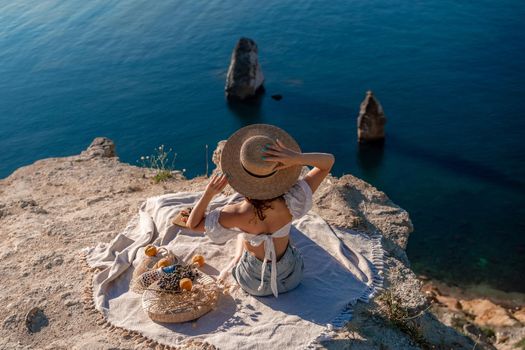 Street photo of a beautiful woman with dark hair in a white top, shorts and a hat having a picnic on a hill overlooking the sea.