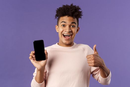 Happy young man, hispanic guy with dreads recommend app, online delivery service or food order, show thumbs-up, smiling excited, holding mobile phone, show smartphone screen.