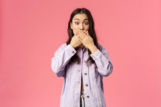 Portrait of surprised young girl found out something she shouldnt have, cover mouth as gasping and staring at camera speechless, trying not slip secret, gossiping, pink background.