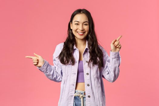 Portrait of stylish assertive cute korean girl giving choice, pointing fingers left and up, promote products, give advice where find useful links on online shops, internet stores, pink background.