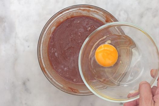 Top view woman hand putting raw egg in glass bowl as ingredient of cake medovik to put it in chocolate batter on marble background