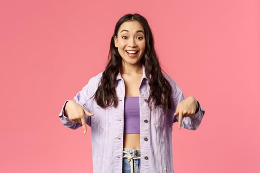 Portrait of excited, happy smiling korean girl in denim jacket, talking about cool new product in store, online shopping site or application, recommend click link, pointing fingers down.