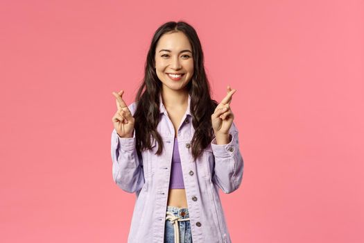 Portrait of excited attractive young korean girl having faith everything be good, keep fingers crossed while making wish, dreaming about desire come true, pink background.