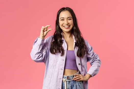 Little effort but big profit. Portrait of cheerful, attractive and carefree modern girl in denim jacket over crop-top, show something is small or tiny, laughing pleased smiling, pink background.