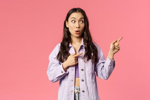 Portrait of enthusiastic, excited pretty asian woman in denim jacket, open mouth in surprise, say wow seeing cool thing, pointing and looking right, standing pink background astounded.