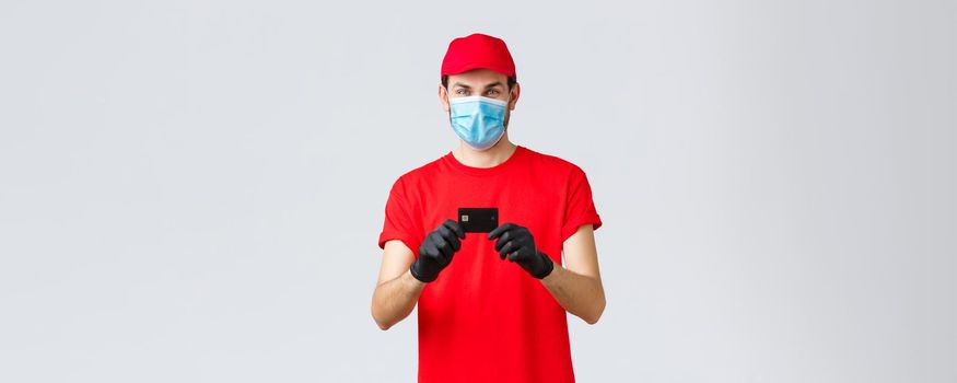 Contactless delivery, payment and online shopping during covid-19, self-quarantine. Cheerful courier in red uniform cap and t-shirt, wear medical face mask, gloves showing credit card, order food.