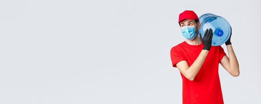 Takeaway, food and groceries delivery, covid-19 contactless orders concept. Carefree courier in red uniform cap, face mask and gloves, hold bottled water on shoulder, look away with smile.