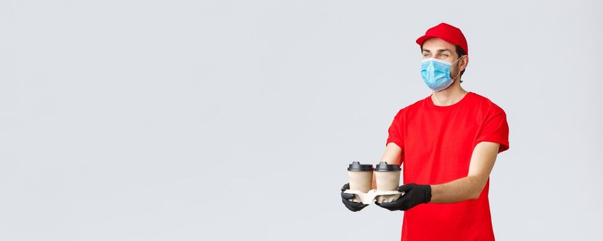 Takeaway, food and groceries delivery, covid-19 contactless orders concept. Pleasant courier in red uniform, face mask and gloves, giving out coffee to customer, stand grey background.