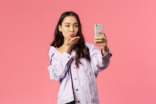 Online lifestyle, people and beauty concept. Lovely modern and stylish asian girl, internet blogger sending kisses via phone camera, taking selfie or record video, pink background.