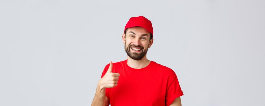 Online shopping, delivery during quarantine and takeaway concept. Smiling handsome bearded delivery guy or courier in red uniform cap, show supportive thumb-up, guarantee safe order transfer.