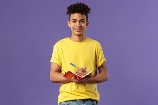 Portrait of young hispanic male student studying online courses, writing down lecture, making personal schedule or taking notes in planner, look camera enthusiastic and interested, purple background