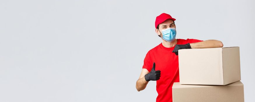 Packages and parcels delivery, covid-19 quarantine and transfer orders. Confident courier in red uniform, gloves and medical mask, encourage call service, show thumb-up lean on boxes.