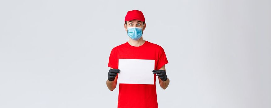 Covid-19, self-quarantine, online shopping and shipping concept. Excited courier in red uniform, gloves and face mask with blank piece paper and stare surprised camera, showing company info or banner.