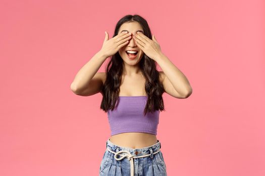 Portrait of excited enthusiastic good-looking woman in crop-top, awaiting for command to open eyes, smiling amused, blindfolded waiting surprise gift, standing pink background.