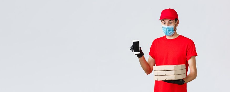 Food delivery, application, online grocery, contactless shopping and covid-19 concept. Amused funny courier in red uniform, face mask and gloves, showing smartphone screen app and hold pizza boxes.