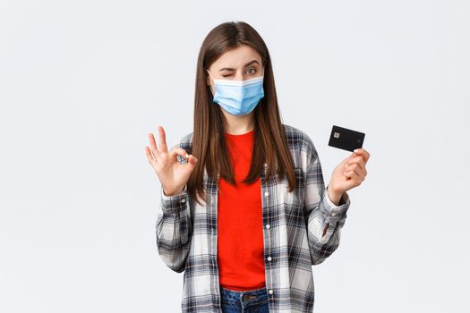 Coronavirus outbreak, working from home, online shopping and contactless payment concept. Pleased young female bank customer in medical mask, show okay sign and credit card, wink.