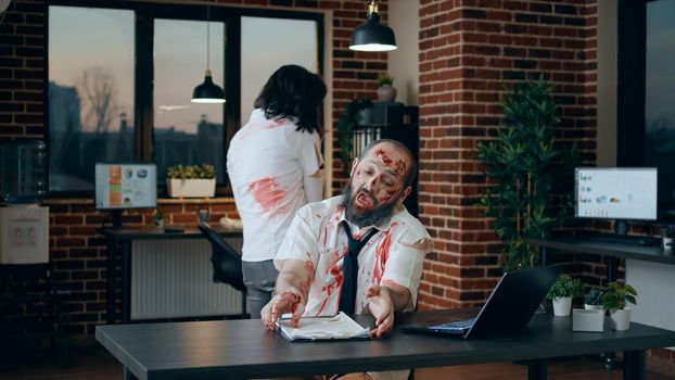 Creepy mindless zombie sitting in office while working on modern laptop. Spooky brain dead monster with deep and bloody scars typing on portable computer while dead walking coworker brings clipboard.