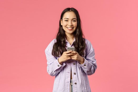 Online lifestyle, people and beauty concept. Cheerful stylish young girl using mobile phone and smiling pleased at camera, chatting during lockdown, use education app, pink background.