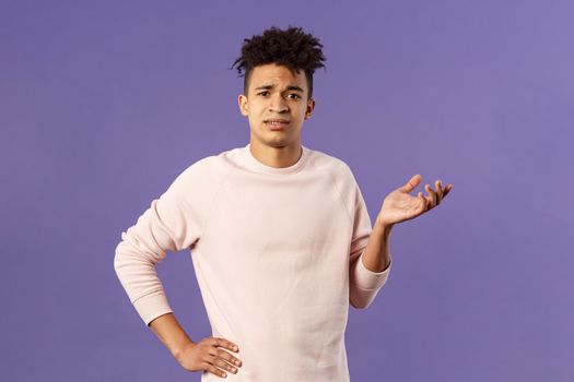 So what, why asking. Portrait of unbothered and careless, ignorant young man dont care on people rules, raising hand in dismay and confusion, being puzzled, look uninterested, purple background.