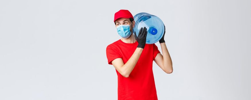 Takeaway, food and groceries delivery, covid-19 contactless orders concept. Carefree courier in red uniform cap, face mask and gloves, hold bottled water on shoulder, look away with smile.