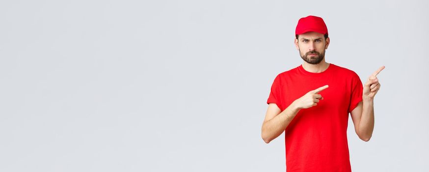 Online shopping, delivery during quarantine and takeaway concept. Displeased angry courier in red uniform cap and t-shirt, frowning grumpy, pointing fingers right in disapproval, feel bothered.