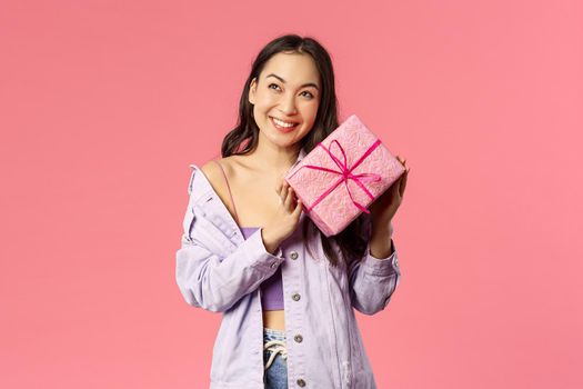 Curious whats inside. Portrait of cute silly asian girl shaking box with gift to guess what is it, smiling amused, leaning to present with ear trying hear movement, standing pink background.