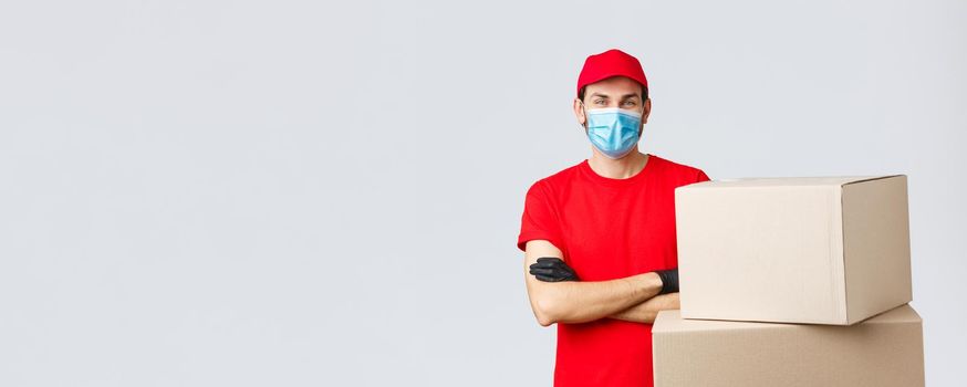 Packages and parcels delivery, covid-19 quarantine and transfer orders. Confident young courier in red uniform, gloves and medical mask, cross arms as standing boxes, grey background.