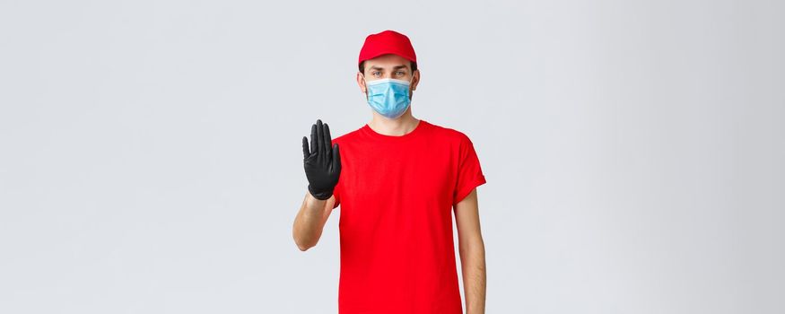 Covid-19, self-quarantine, online shopping concept. Confident delivery man in red uniform, gloves and face mask, raise hand in stop sign, prevent client step to storage without protective equipment.